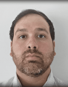 Professional headshot of a middle-aged man with dark hair and graying beard wearing a light gray collared shirt against a white background