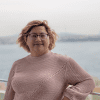 Portrait of a smiling person wearing round glasses and a pink knitted sweater, posing outdoors by a waterfront with a scenic lake background.